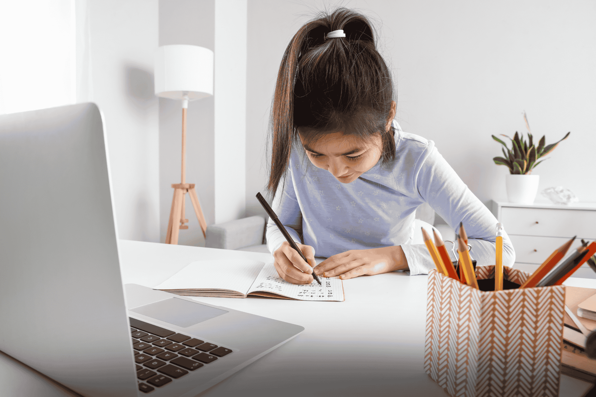 A young girl writes in a notebook at a desk with a laptop and pencils nearby.