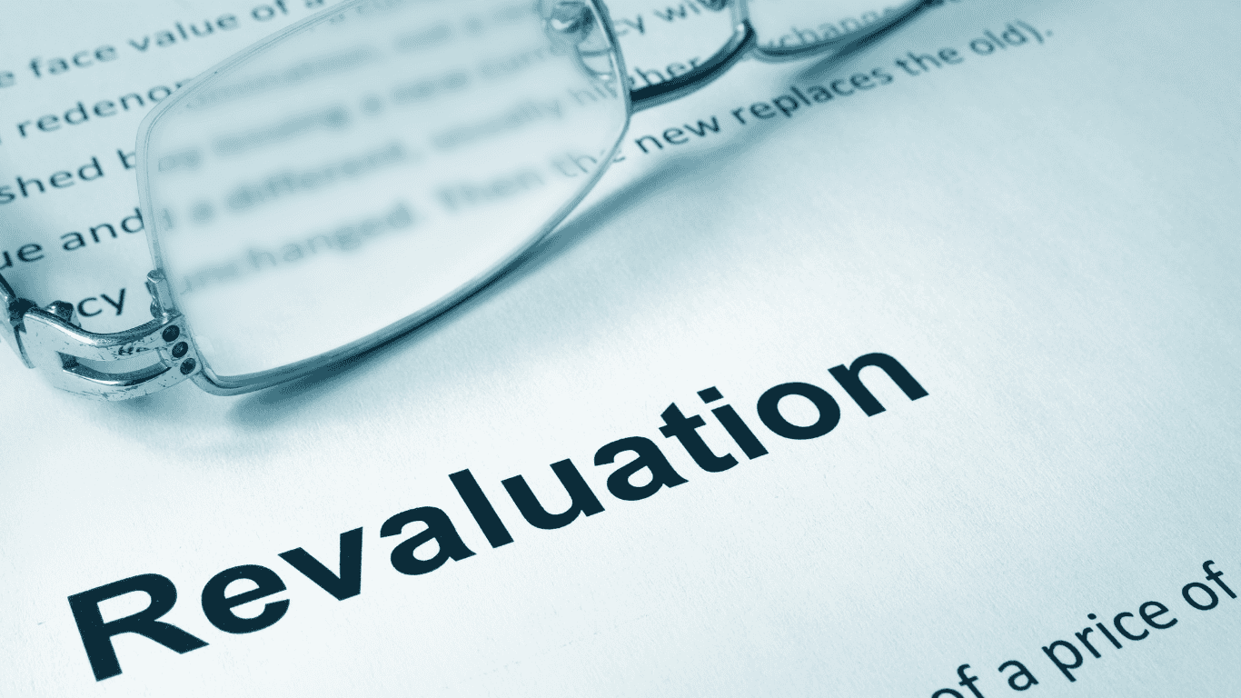 A close-up of the word 'Revaluation' printed on a document, with a pair of reading glasses partially covering the text. The image has a blue tint, and the background text is blurred.