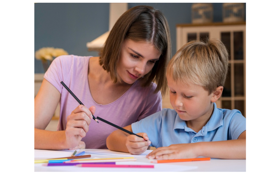 A young woman helps a little boy with his drawing, guiding his hand with a pencil. They are sitting at a table with colorful pencils and paper, engaged in a learning activity.