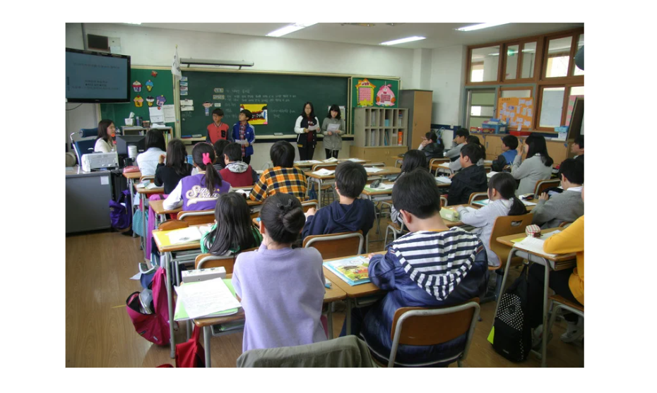 Students sit at their desks, attentively watching a group presentation at the front. A teacher observes from the side, while a blackboard filled with educational materials serves as the backdrop.